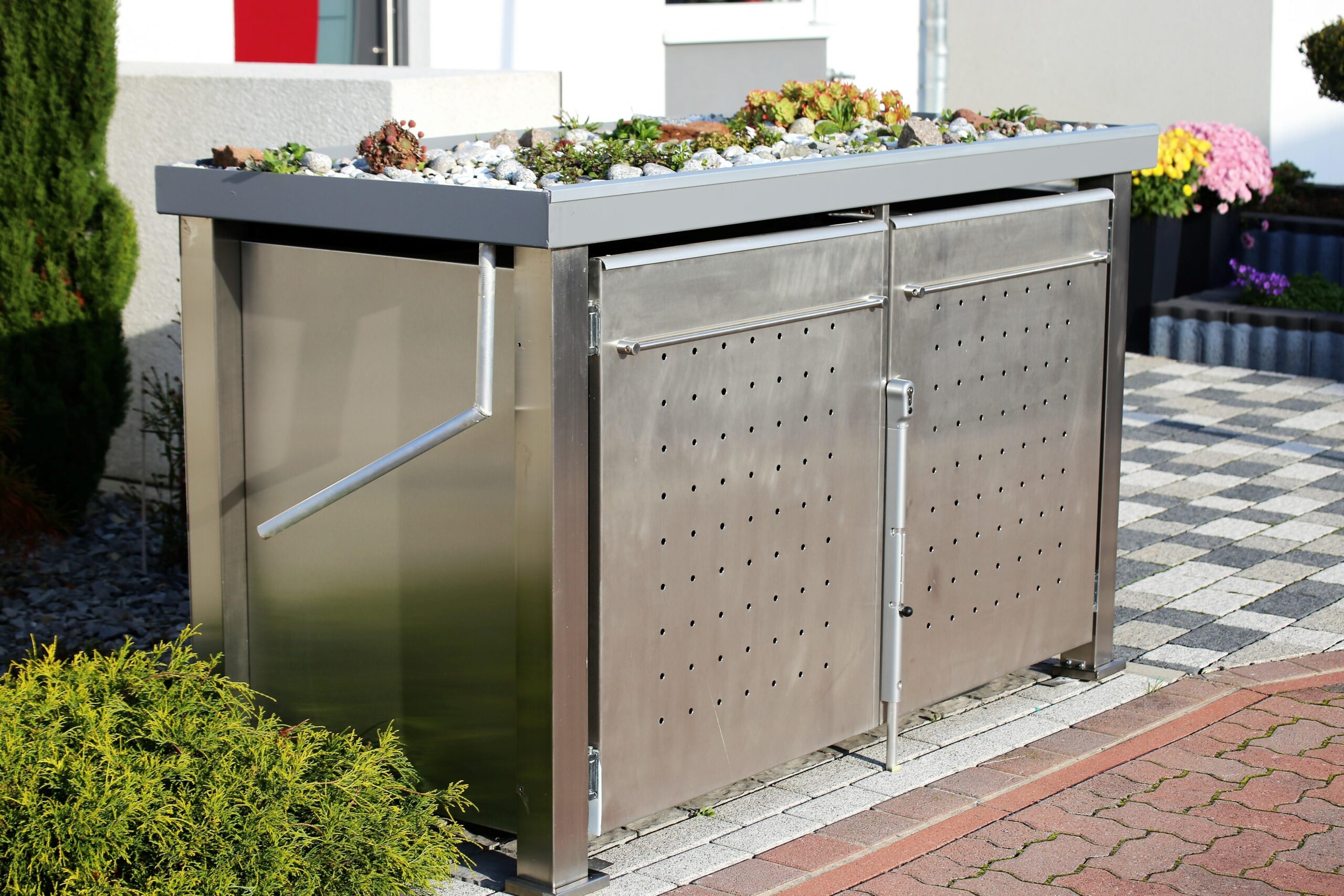 Eine elegante Mülltonnenbox aus Edelstahl mit bepflanztem Dach, steht auf einem gepflasterten Bereich in einem gepflegten Garten vor einem Haus.