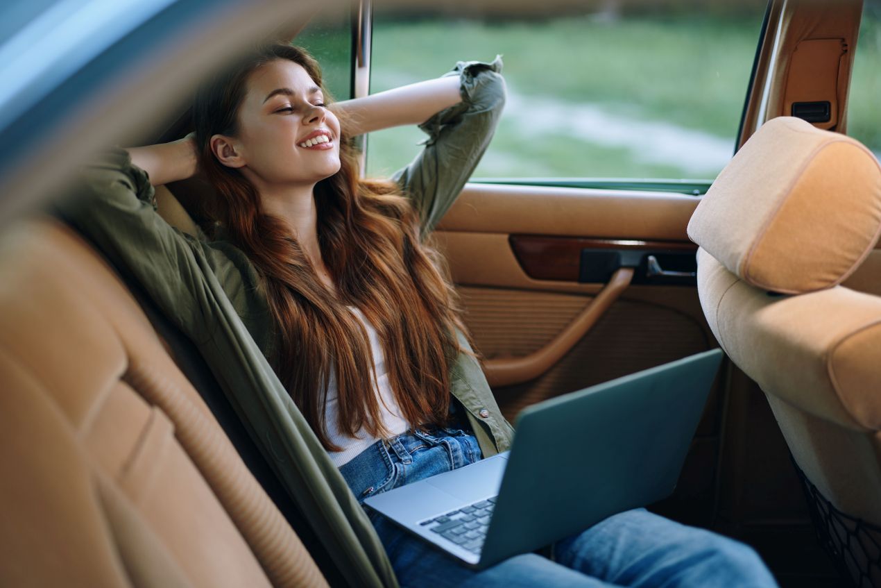 Junge Frau lehnt sich entspannt im Auto zurück, hält einen Laptop auf dem Schoß und genießt flexible Arbeitsbedingungen.