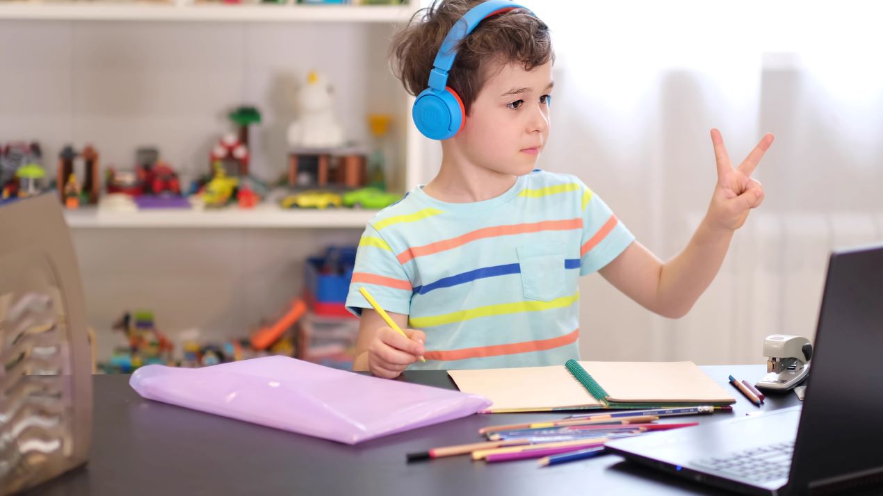 Ein Junge mit blauen Kopfhörern lernt mit Mathe Nachhilfe Online. Er zeigt zwei Finger und arbeitet konzentriert an seinen Aufgaben mit Laptop und Schreibmaterial.