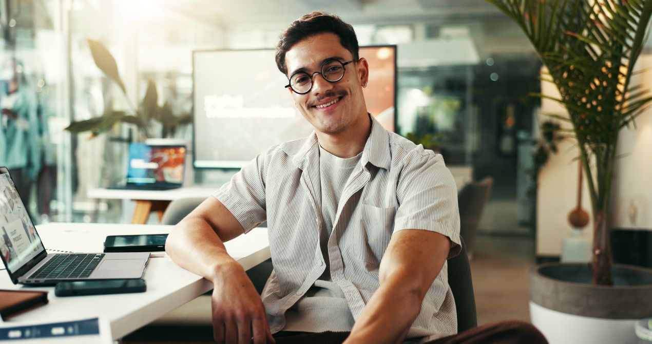 Junger Mann mit Brille und Laptop lächelt in modernem Büro – klare, fehlerfreie Texte dank professionellem Lektorat.